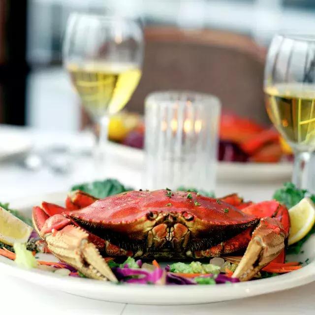 在一家餐馆里，一只邓杰尼斯蟹坐在盘子里，背景是两杯白葡萄酒.